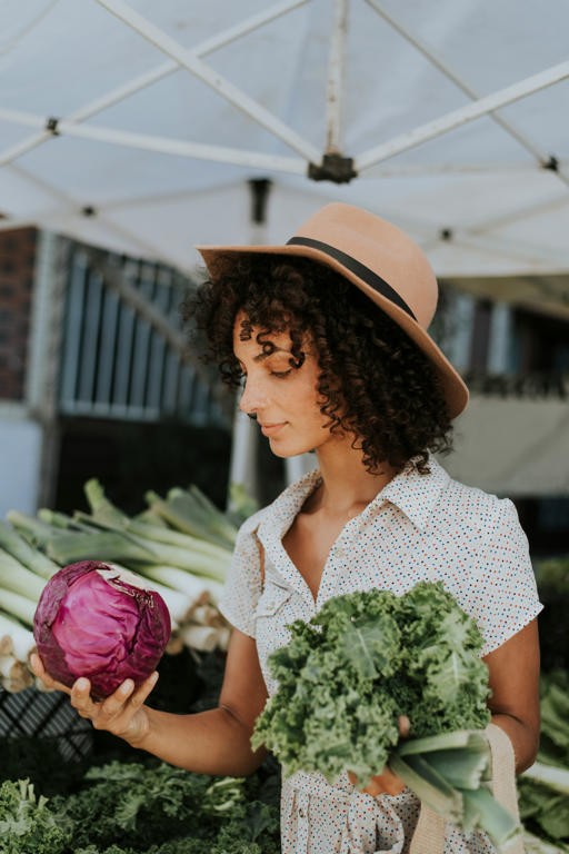 A woman holds kale in one hand and cabbage in the other at a farmers market. She inspects the cabbage to see if it's fresh.